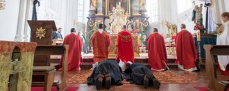 in der Kirche: Pater in rot knien vor dem Altar - dahinter liegen schwarz gekleidete Pater 